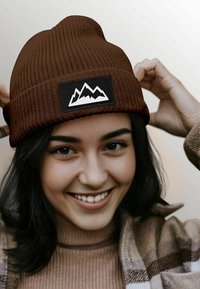 Brown knitted beanie cap with a black patch featuring white mountain graphics, tightly fitted at the top, worn by a person smiling.