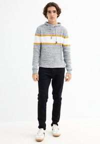 Sudadera de hilo gris con rayas amarillas y blancas, tela texturizada, capucha con cordones, combinada con pantalones negros y zapatillas blancas.
