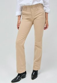 Pantalones beige de corte slim con dos bolsillos frontales. Cuentan con cierre de botón y cremallera, combinados con botas negras de tacón.