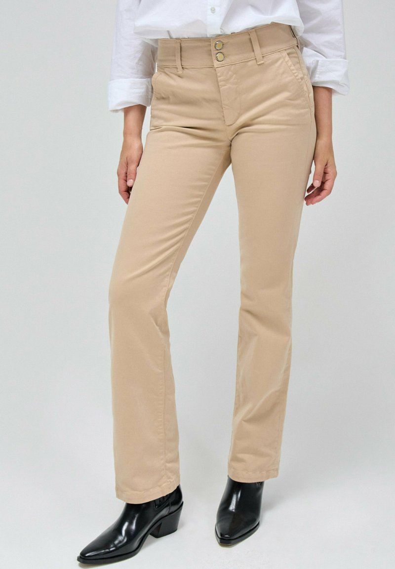 Pantalones beige de corte slim con dos bolsillos frontales. Cuentan con cierre de botón y cremallera, combinados con botas negras de tacón.