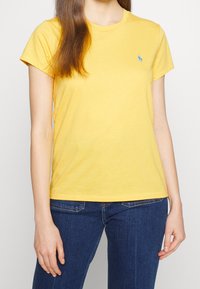 Gul bomullströja med kort ärm, rund halsringning och en liten blå broderad logotyp på vänster bröst. Matchad med blå jeans.