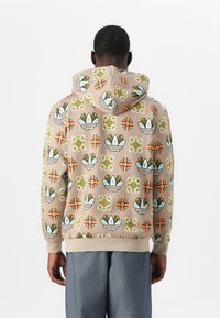 Uomo che indossa un hoodie beige con motivi floreali geometrici verdi, arancioni e bianchi, abbinato a pantaloni blu larghi, di spalle.