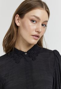 Blusa nera con colletto in pizzo con ricami intricati, chiusura frontale con bottoni e tessuto strutturato con maniche a sbuffo.