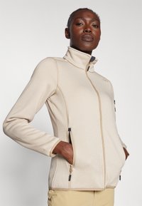 Veste zippée beige en tissu texturé, col montant et coupe ajustée. Dotée de poches latérales zippées et d'accents contrastés.