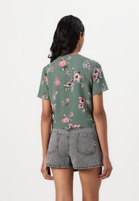 Top corto a maniche corte verde floreale con fiori rosa e blu, abbinato a pantaloni corti in denim grigi con tasche posteriori e orlo sfrangiato.