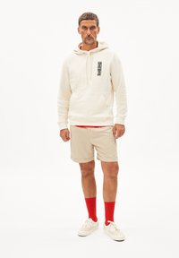 Felpa crema con grafica nera, pantaloni corti beige, calzini rossi e sneakers chiare. Il tessuto sembra morbido con una vestibilità comoda e un design informale.