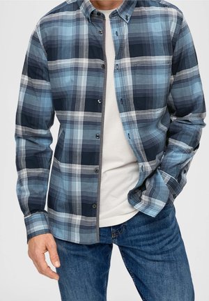 Mann trägt ein blau-grau kariertes Hemd über einem weißen T-Shirt mit blauen Jeans, Hände in den Taschen und entspannte Haltung.