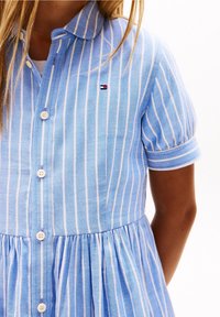 Abito a righe azzurro chiaro con maniche corte, chiusura frontale con bottoni, gonna plissettata e piccolo logo sul petto. Realizzato in tessuto intrecciato.