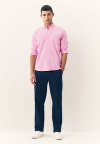 Light pink button-up shirt with rolled sleeves, paired with navy pants and white sneakers, featuring a small logo on the shirt's chest.