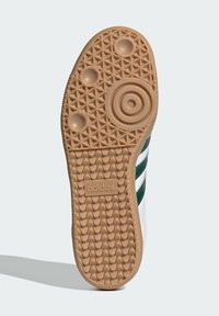 Bijela potplat tenisice s teksturiranom gumom, koji sadrži valoviti uzorak, kružne naglaske i istaknuti "adidas" logo u boji smeđa.