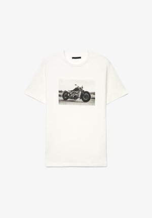 Weißes T-Shirt mit einem schwarz-weißen Foto eines alten Motorrads, das auf einer unbefestigten Straße geparkt ist, auf der Vorderseite gedruckt.