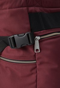 Bordeaux stoffpose med en teksturert overflate, med en svart plastspenne og en sølvglitrende glidelås med en taustropp. Detaljene er i svart nylon.