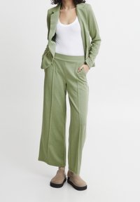Zielona dopasowana marynarka i szerokie spodnie z miękkiego materiału, z kieszeniami i luźnym krojem, w zestawieniu z białym topem na ramiączkach i beżowymi butami.