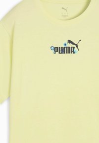 Vaaleankeltainen puuvillapaita, jossa on musta "PUMA" -logo, sinisiä kukkakirjontoja ja musta pantterin siluetti. Pyöreä pääntie.
