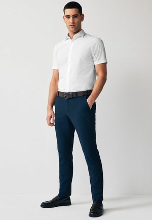 Bijela košulja kratkih rukava s gumbima, mornarsko plave hlače, crni remen i mokasine. Odjevna kombinacija karakterizira elegantan, prilagođen kroj i glatke teksture.