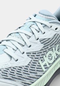 Kengät urheiluun vaaleansinisellä värillä, verkkopintalla, harmailla teksturoiduilla sivuilla ja valkoisilla nauhoilla. Varustettu "Hoka" -brändillä ja "Fate Speed" -tekstillä kielekkeessä.