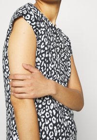 Kvinna som bär en kortärmad topp med svart och vitt leopardmönster, med en arm som håller den andra strax ovanför armb ågen mot en vit bakgrund.