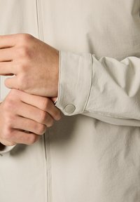 Lichtbeige jaszoom met een knoopsluiting, voorzien van een gladde textuur en zichtbare stiksel details over de stof.