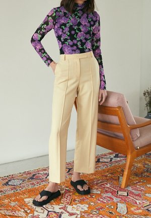 Femme portant un pantalon beige taille haute, un haut à manches longues noir et violet à motifs floraux et des sandales noires, debout sur un tapis à motifs à côté d'une chaise en bois.