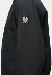 Μανίκι μαύρου μπουφάν με μπεζ τιμή Belstaff που φέρει ένα φτερωτό έμβλημα και κείμενο στο πάνω μέρος του βραχίονα.