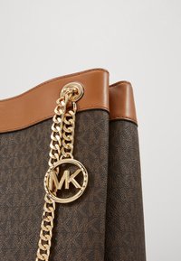 Gros plan d'un coin d'un sac à main Michael Kors marron avec une bandoulière chaîne dorée et un pendentif logo MK sur un matériau monogramme texturé.