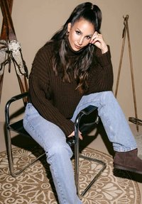 Bruine geribde trui, lichtblauwe jeans en donkerbruine enkellaarzen; zittend op een chroomstoel met een gemêleerd vloerkleed eronder.