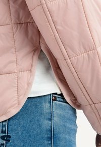 Lys pink quilted jakke med en glat finish, med en rund kant og diskrete syninger, båret over en hvid skjorte og blå jeans.