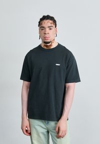 Black cotton t-shirt with short sleeves and a crew neckline, featuring a white "OBEY" logo on the chest, worn with light blue jeans.