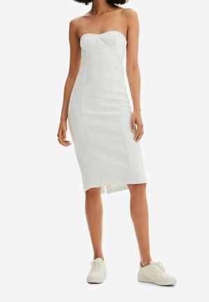 Frau trägt ein trägerloses weißes Midikleid mit kleinem Frontschlitz, kombiniert mit weißen Sneakern, steht vor einem schlichten Hintergrund.