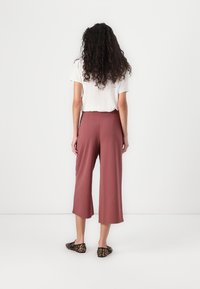 Femme aux longs cheveux bouclés portant un haut blanc à manches courtes, un pantalon large cropped mauve et des ballerines à imprimé léopard, faisant face à un mur blanc.