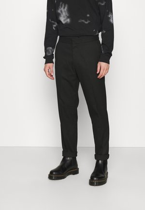 Person iført sorte bukser med opbuk ved anklerne, sorte læderstøvler med gul syning og en sort batiklangærmet skjorte, stående mod en hvid væg.