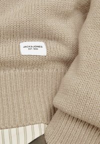 Pull en tricot beige avec des poignets côtelés, arborant une étiquette blanche avec le texte noir « JACK & JONES EST. 1990 » et une doublure intérieure rayée.