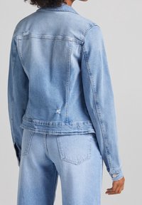 En person klädd i en ljusblå denimjacka och matchande jeans, sedd bakifrån mot en enfärgad grå bakgrund.