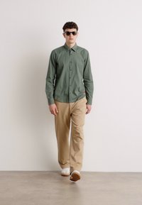 Cămașă verde cu butoni, pantaloni bej și adidași albi. Modelul poartă ochelari de soare, stând în fața unui fundal neutru.