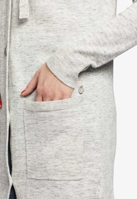 Main avec des ongles peints en rouge reposant dans la poche avant d'un cardigan en maille grise clair.