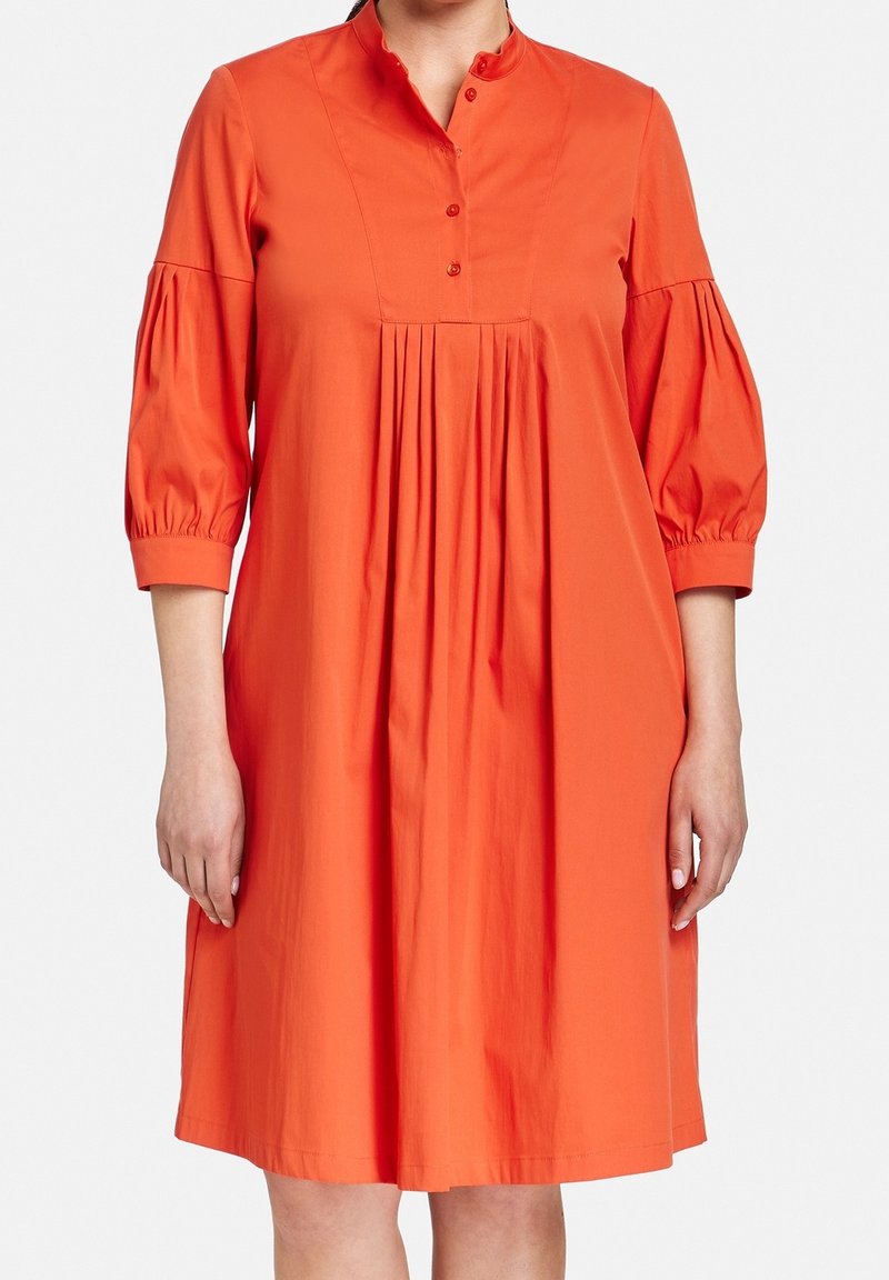 Vestito di cotone arancione con colletto alla mandarina con bottoni, maniche a sbuffo 3/4 e un design frontale plissettato. Texture liscia, silhouette alla lunghezza del ginocchio.