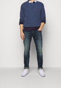 Man i en marinblå sweatshirt över en vit skjorta med krage, blekblå jeans med uppvikta ben och vita sneakers som står mot en enfärgad bakgrund.
