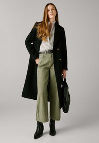 Cappotto di lana nero, blusa bianca con texture, pantaloni a palazzo verde oliva, cintura nera con fibbia, stivali neri con tacco, in mano una borsa nera.