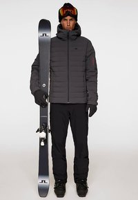 Skidutrustning inkluderar en svart vadderad jacka, svarta handskar och svarta byxor. Personen håller en svart skida med vita bindningar.