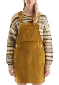 Kordon overall jurk in mosterdgeel met verstelbare schouderbanden, een voorzak en zijknopen als accenten, gedragen over een gestreepte gebreide trui.