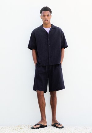 Jeune homme debout contre un mur blanc portant une chemise noire à manches courtes avec boutons, un short assorti et des tongs noires, les mains dans les poches.