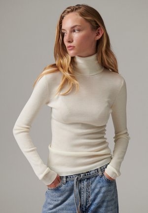 Jeune femme aux longs cheveux blonds portant un pull à col roulé crème ajusté et un jean bleu, les mains dans les poches, regardant sur le côté.