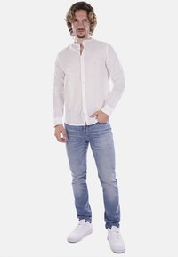 Camicia bianca a maniche lunghe con patta bottonata, realizzata in tessuto leggero, abbinata a jeans azzurri e sneakers bianche.
