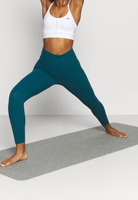 Tealfärgade leggings och en vit sport-bh på en person i en krigare-yogaposition, stående på en grå matta, som visar upp en åtsittande design.