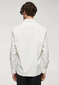 Homme aux cheveux bouclés portant des lunettes de soleil noires et une chemise blanche à manches longues à rayures verticales, vu de dos.