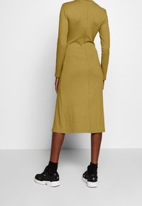 Olivgrünes, geripptes Midikleid mit langen Ärmeln, taillierter Silhouette und rückseitigen Nahtdetails. Kombiniert mit schwarzen Sportschuhen.