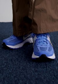 Ténis atléticos Nike em azul claro com painéis em malha, superfícies texturizadas, detalhes em preto e sola branca, combinados com calças castanhas.