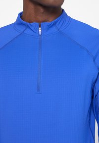 Niebieski półzapínany polar wykonany z teksturowanej tkaniny, z kołnierzem stójkowym i kontrastowymi szwami wzmacniającymi.