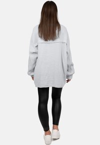 Maglia oversize grigio chiaro con texture e maniche lunghe, indossata con leggings neri e sneaker bianche, vista da dietro.