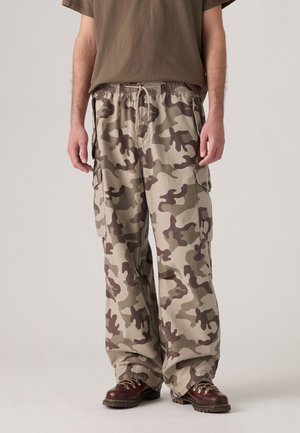 Man wearing loose beige and brown camouflage cargo pants with pockets, brown boots, and a brown t-shirt, standing against a plain backdrop.